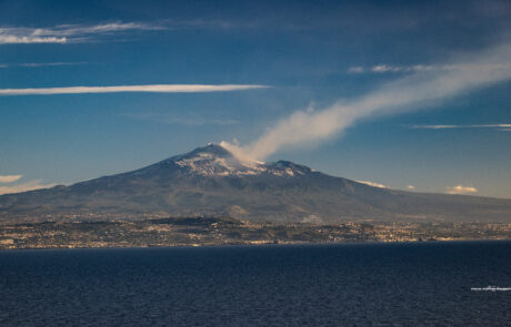 Mount Etna volcano smoking under a hazy sky, seen from the coast