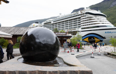 Cruise ship AIDAprima docked at Flam harbor with a black sphere sculpture and tourists nearby