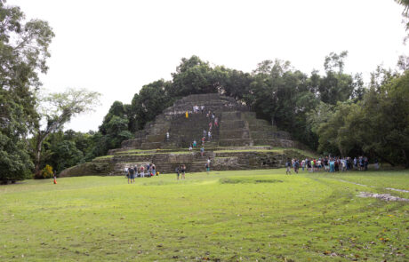 Wide view of a grass-covered Mayan pyramid with visitors, Lamanai